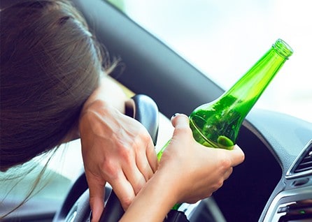 Person resting head on steering wheel while holding a green bottle.