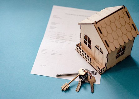 A small wooden house model sits on a document. Three keys on a keyring are placed nearby on a blue surface.