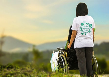 A person stands beside a wheelchair, facing a scenic landscape with hills and a clear sky. The person's shirt displays an Earth-themed design.