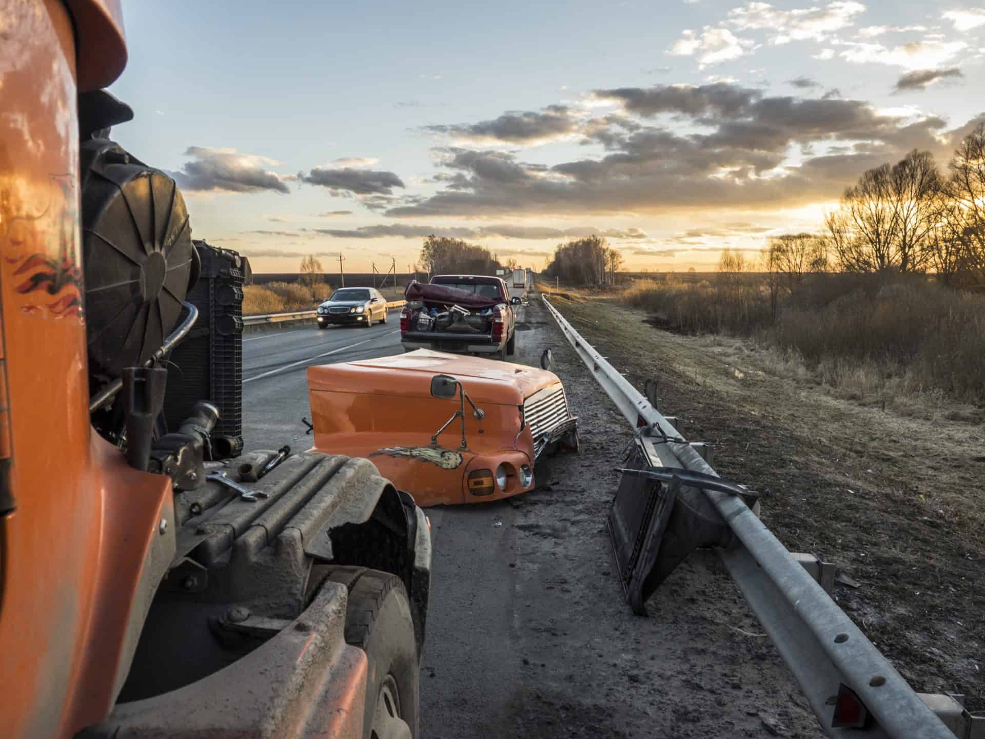 Two vehicles involved in an accident on a highway at sunset, with debris on the road. A semi-truck's front hood is detached, and a car is visibly damaged. Traffic is passing by.